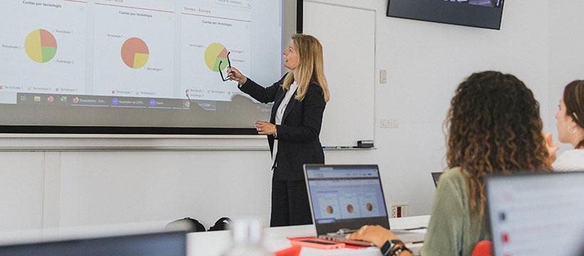 Professor pointing to a pie-chart slide while students with laptops follow along in a bright classroom.