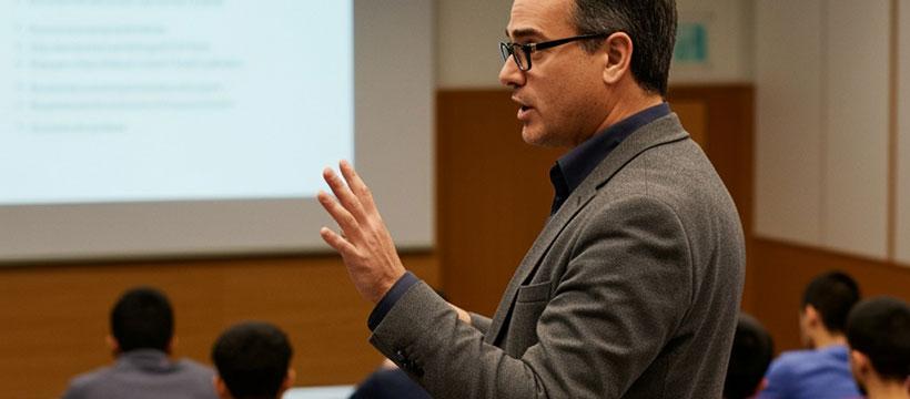 Professor speaking to a class, gesturing during a lecture with a projected slide.